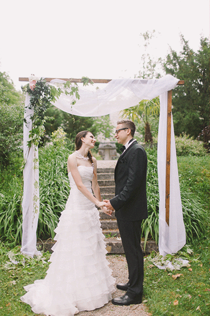 Mladoporočenca stojita pod lesenim obokom, ki je primeren za intimne poroke v vrtu gradu Brdo. Poročni obok je okrašen s cvetjem in blagom. Foto: Nastja Kovačič Photography