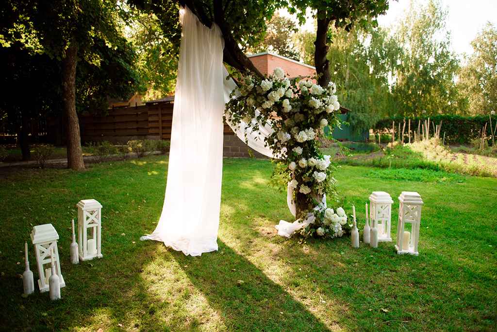 Poroči obred na domačem vrtu pri drevesu, ki je okrašen s cvetjem in belim blagom. Foto: Storija weddings