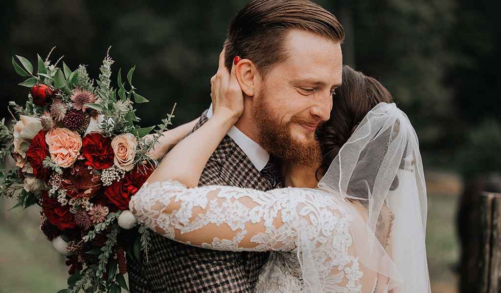 Nevesta v čipkasti poročni obleki z dolgimi rokavi drži poročni šopek v bordo rdeči barvi in ženin v po meri narejeni poročni obleki objeta na posestvu Pule. Foto: Ana Gregorič Photography