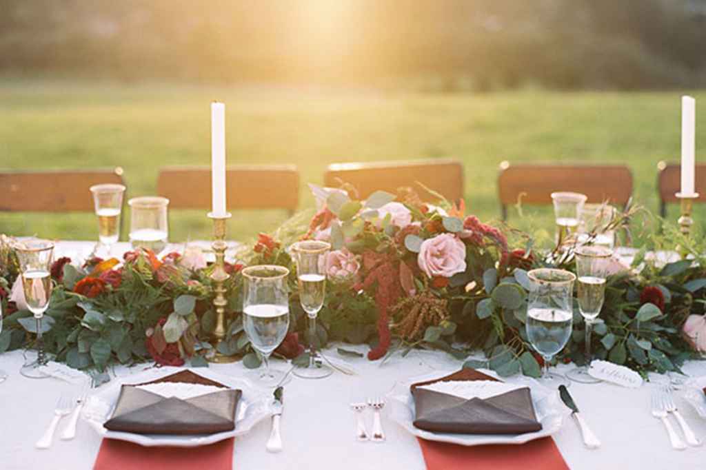 Poročna miza je sestavljena iz belega prta, cvetlične girlande po sredini, kozarci z zlatim robom, zlatimi svečniki, rdečim prtičkom pod krožnikom in na krožniku je poročni meni v rjavi kuverti. Foto: Danielle Poff