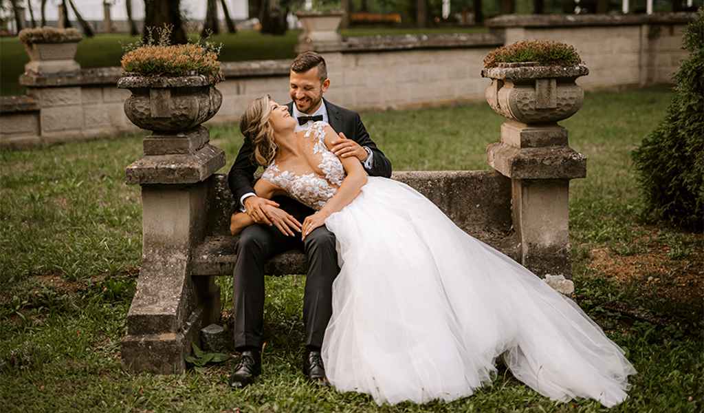 Ženin v prečudoviti črni moški obleki iz salona Morro sedi na kamniti klopci v parku in v rokah drži nevesto v prelepi beli čipkasti poročni obleki. Foto: Ana Gregorič Photography